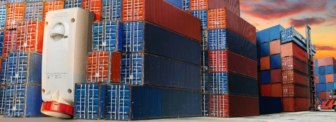 Scame interlocked industrial plug for maritime containers. Detail of a white industrial outlet with red cap, mounted laterally on a container, in a context of port storage with numerous stacked containers of various colors.