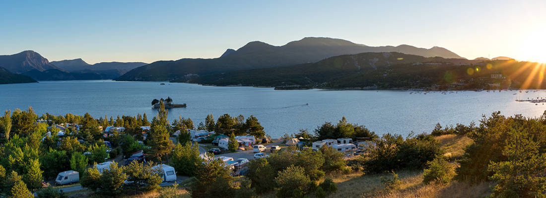 Panoramic view of a campsite with tents and campers surrounded by greenery, overlooking the lake, with mountains in the background and the sun setting on the horizon.